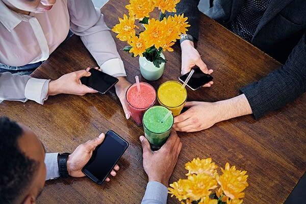 Office group of four people seated at a table with drinks and mobile phones, coordinating a smoothie or juice catering or delivery order