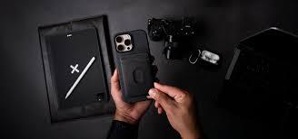 Persona sosteniendo un iPhone 15 Pro Max con funda protectora negra, acompañado de accesorios tecnológicos en un escritorio negro en Pereira, Colombia.