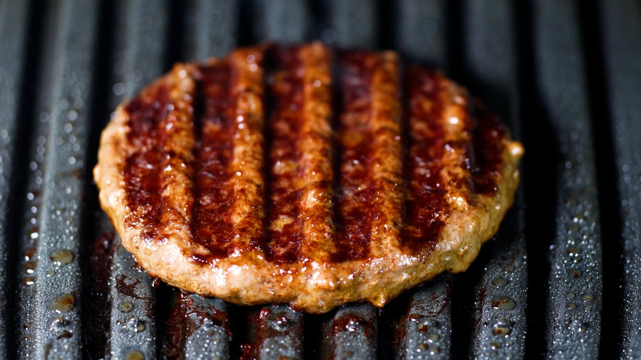 Close-up of a juicy beef burger patty cooking on a ridged grill pan, showing deep char-mark sear lines and glistening juices against the textured black metal surface.