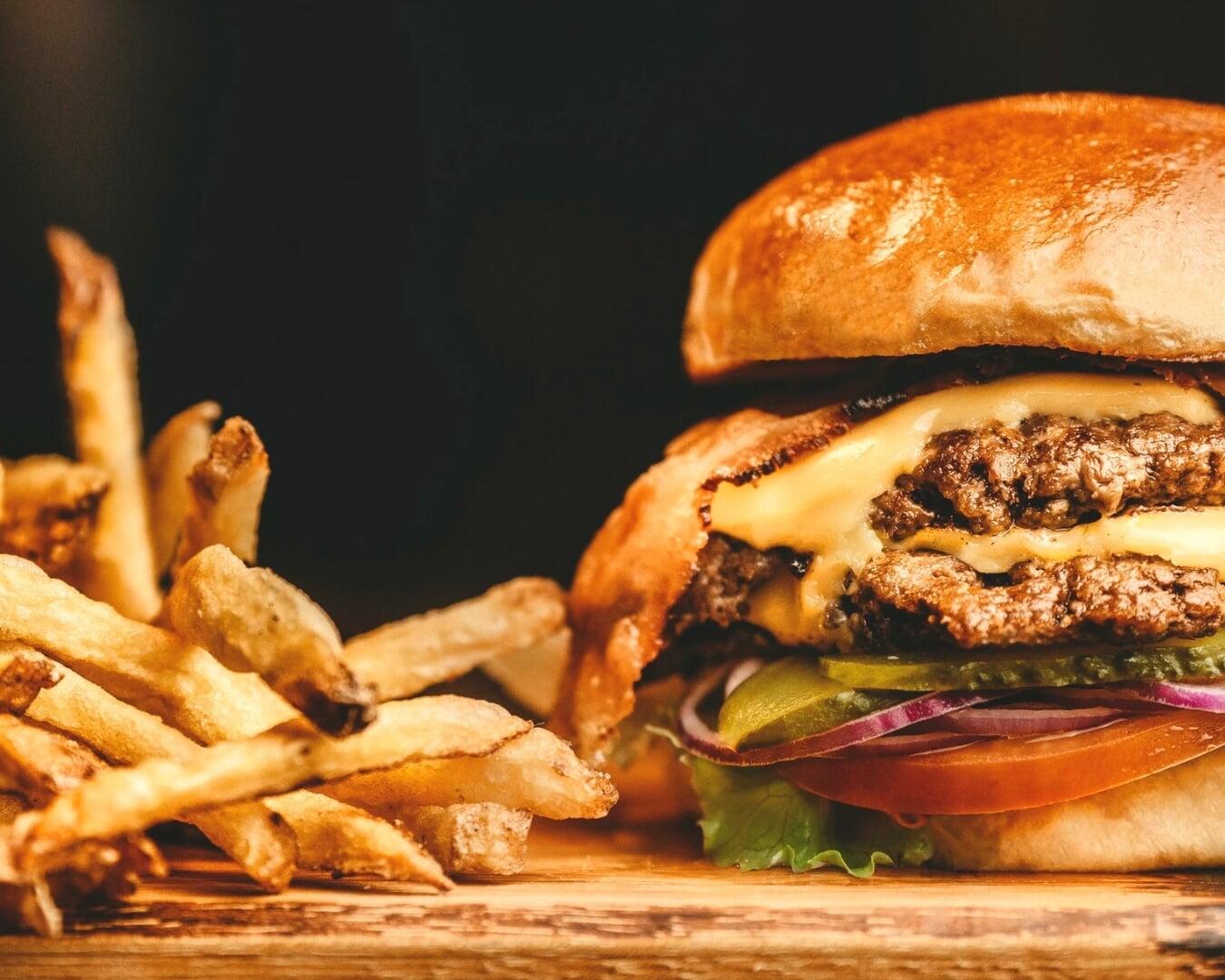 A close-up view of a double cheeseburger—two beef patties with melted cheese, lettuce, tomato, pickles, and a toasted bun—sitting on a wooden surface alongside a pile of golden, crispy French fries against a dark background.