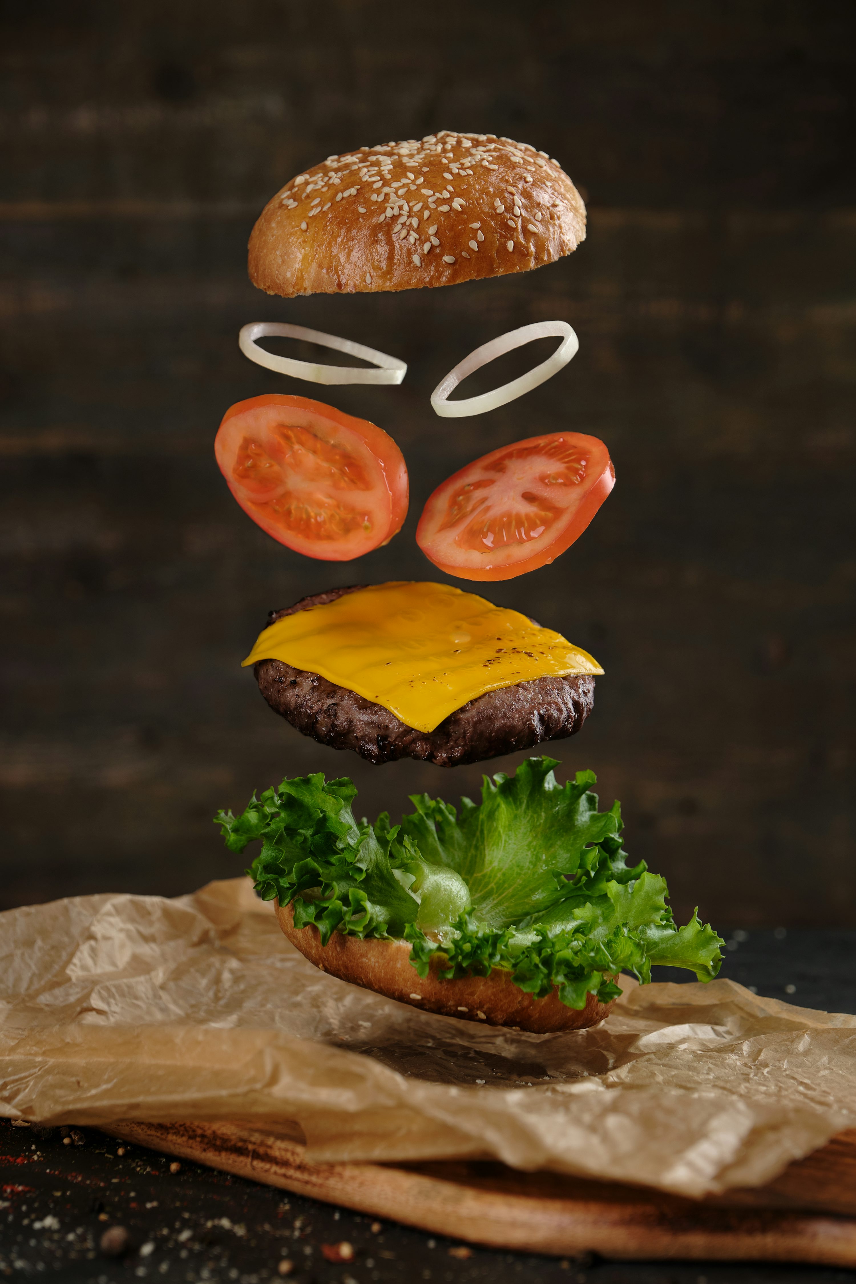 Exploded-view photograph of a cheeseburger hovering above a wooden board: a sesame-seed top bun floats above thin onion rings, tomato slices, a melted-cheddar-topped beef patty, curly green lettuce, and a bottom bun resting on crumpled parchment paper against a dark wood background.
